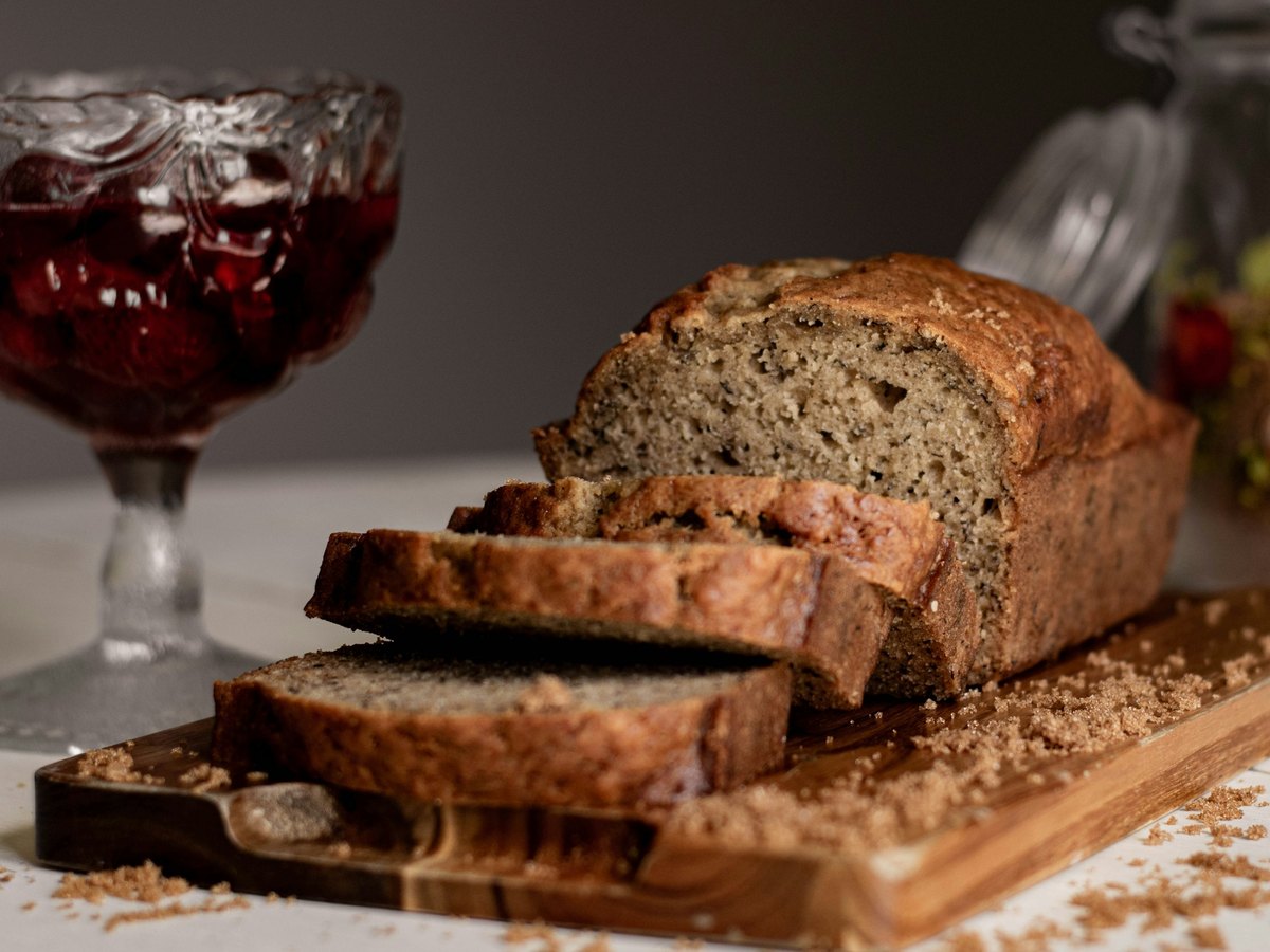 A sliced loaf of banana bread on a wooden board, its top domed and cracked and its crumb dense and flecked, with brown sugar scattered around it