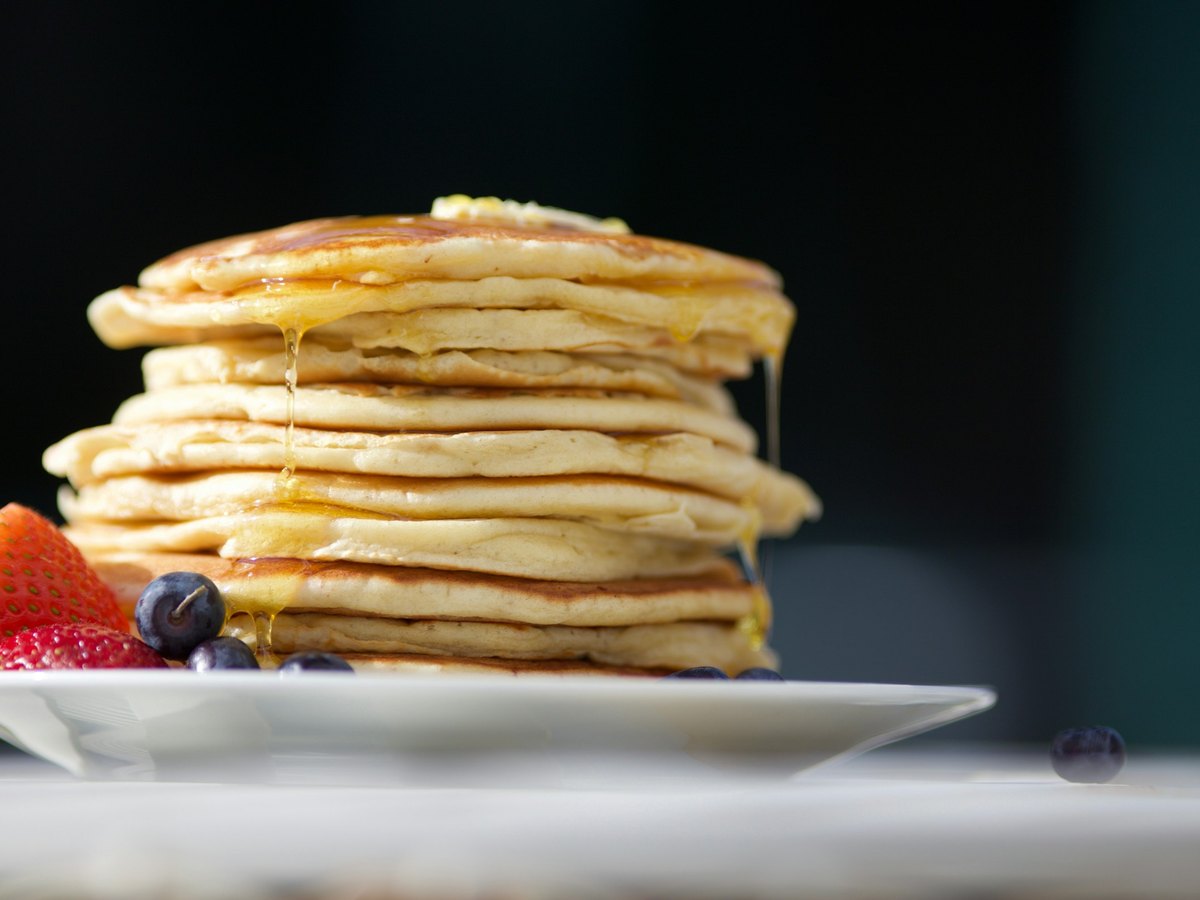 A tall stack of thick pale-golden buttermilk pancakes on a white plate, butter melting on top and syrup running down the sides, with strawberries and blueberries beside them against a dark background