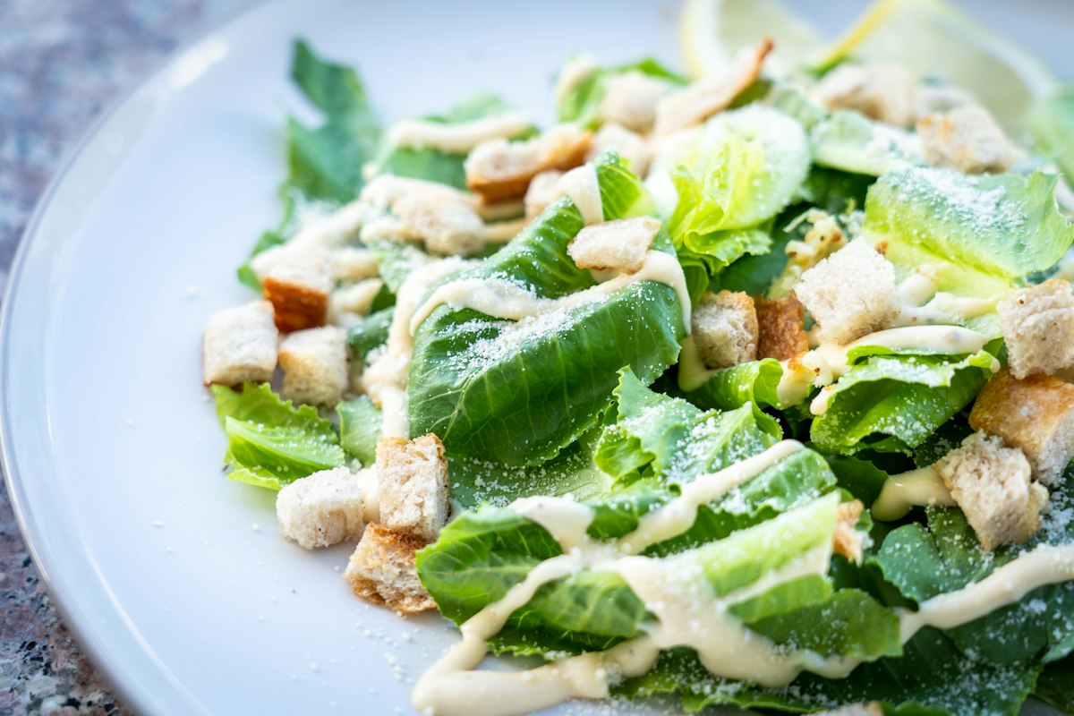 Close-up of a Caesar salad with torn romaine lettuce, golden croutons, shredded parmesan and creamy dressing on a white plate, with a lemon wedge in the background