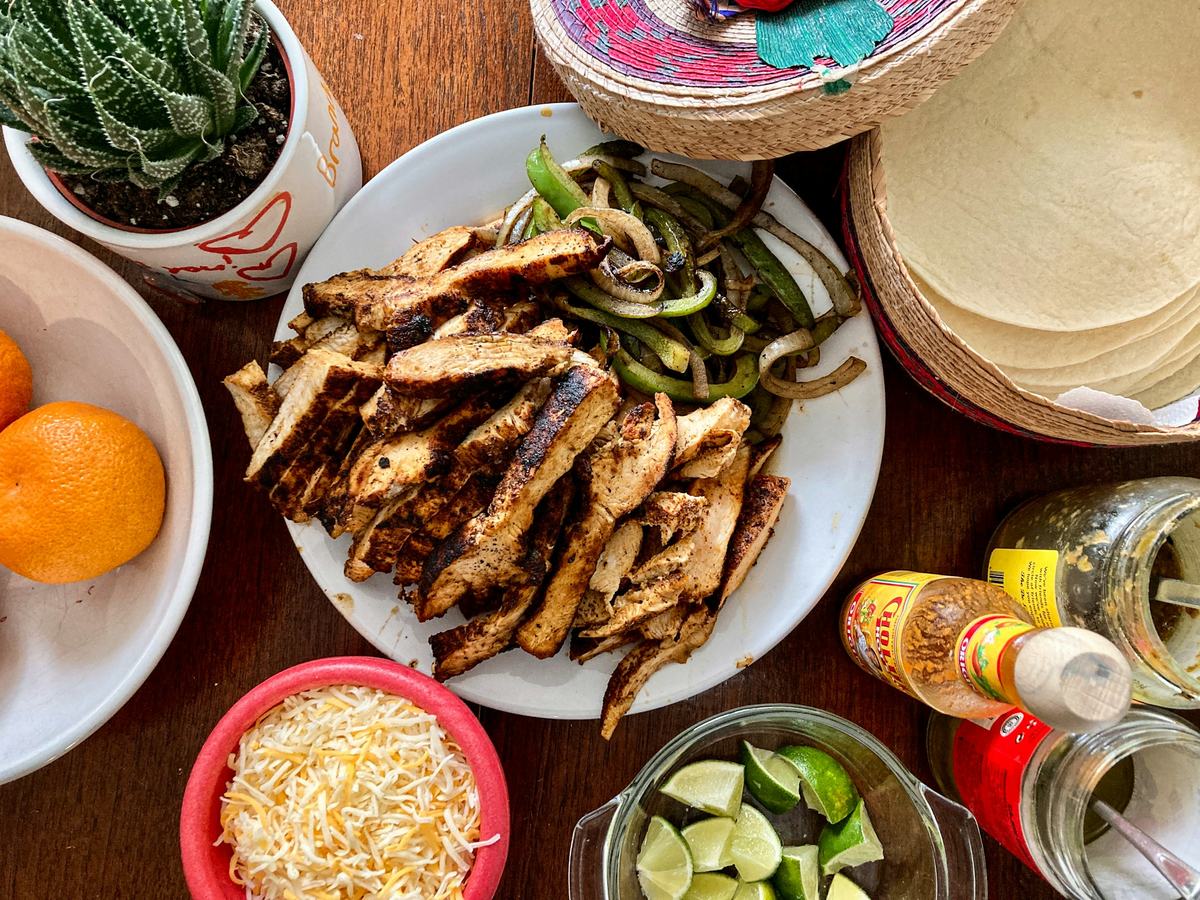 A table from above: a white plate of sliced grilled chicken with charred green peppers and onions, a stack of warm flour tortillas in a woven basket, a bowl of grated cheese, lime wedges and hot sauce