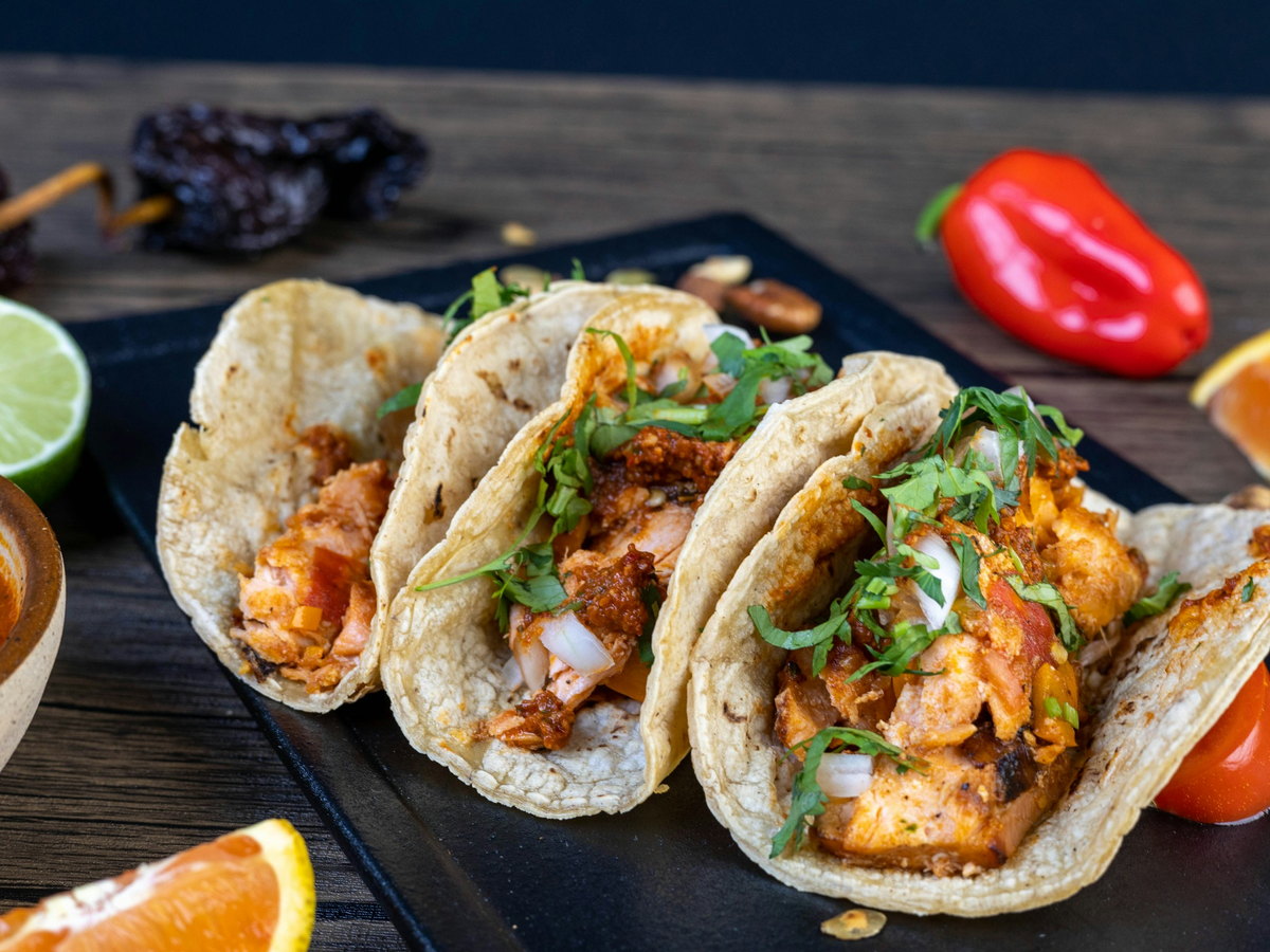 Three soft corn tortillas lined up on a dark slate board, each holding chunks of chipotle-marinated salmon with finely chopped white onion and coriander, a halved lime, dried chillies and orange wedges around them