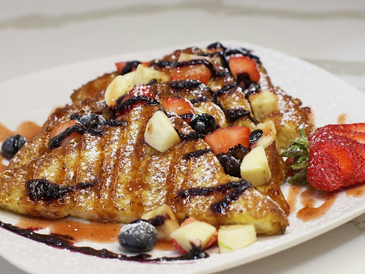 Thick golden-brown slices of French toast on a white plate, dusted with icing sugar and topped with strawberries, blueberries, banana and a dark berry syrup