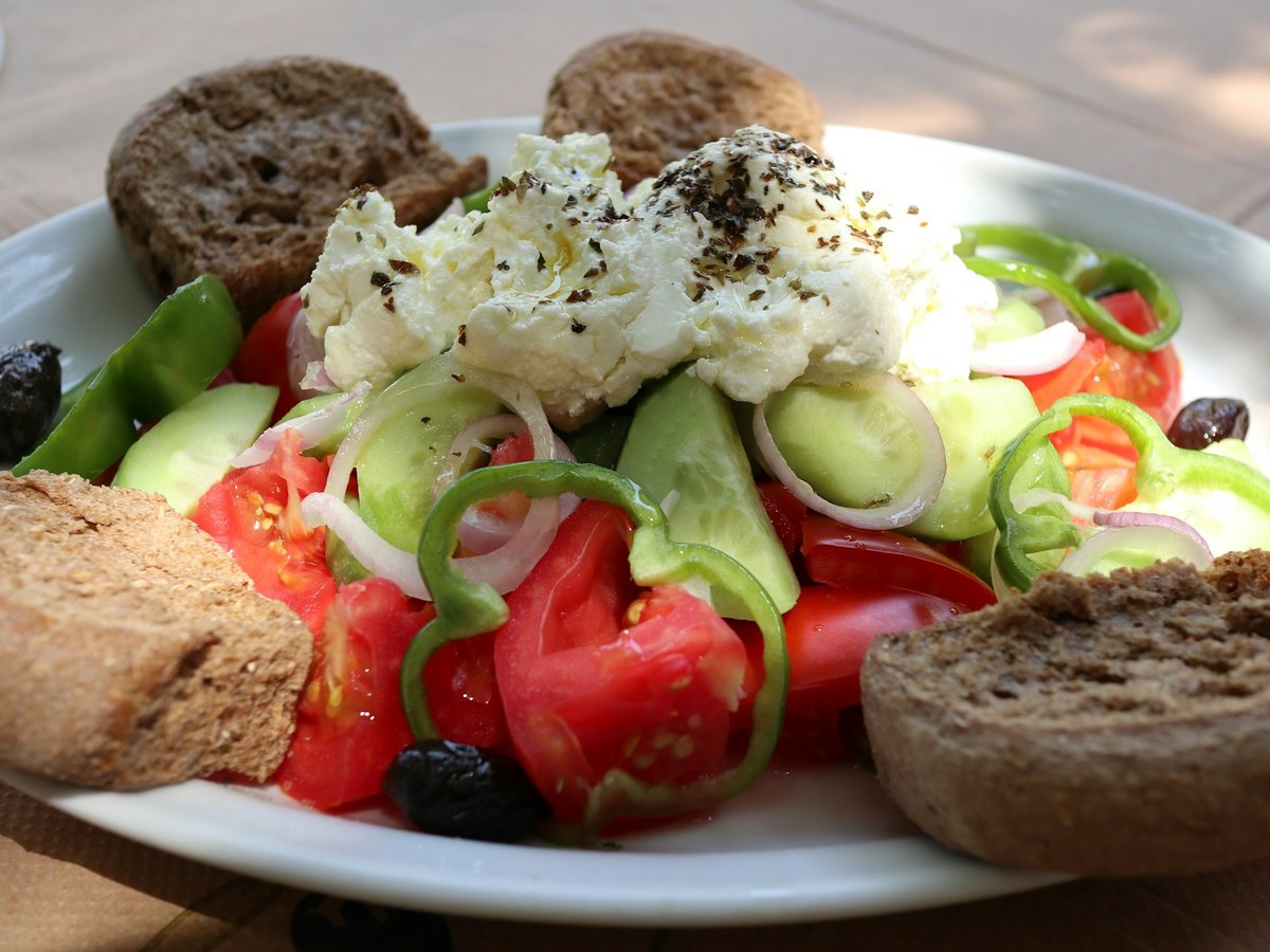 A Greek salad of tomato, cucumber, green pepper, red onion and black olives under a slab of feta, with barley rusks around the plate