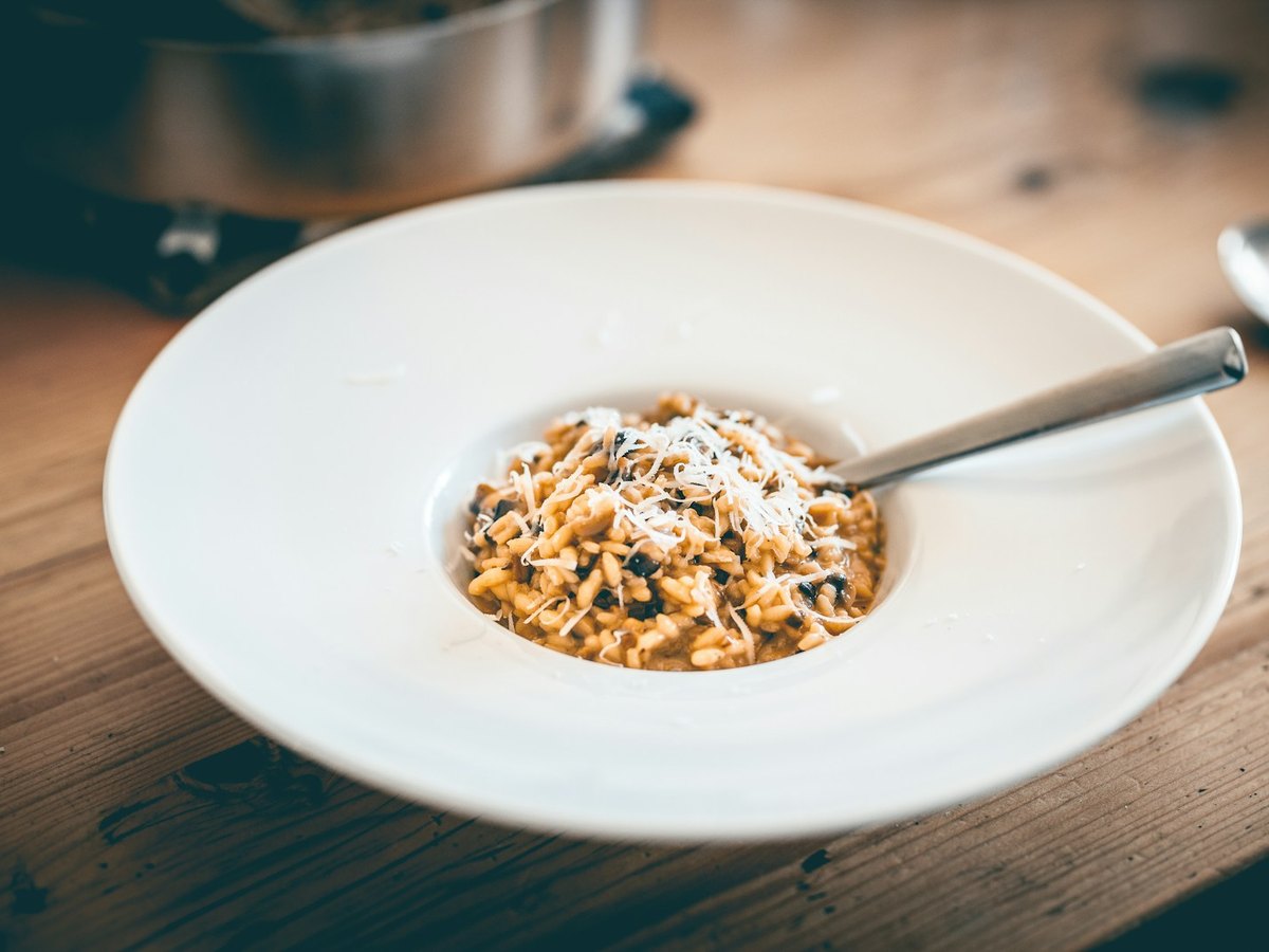A shallow white bowl on a wooden table holding creamy mushroom risotto with dark pieces of mushroom through it, coarsely grated parmesan on top and a spoon resting in the bowl, with the pan it was cooked in out of focus behind