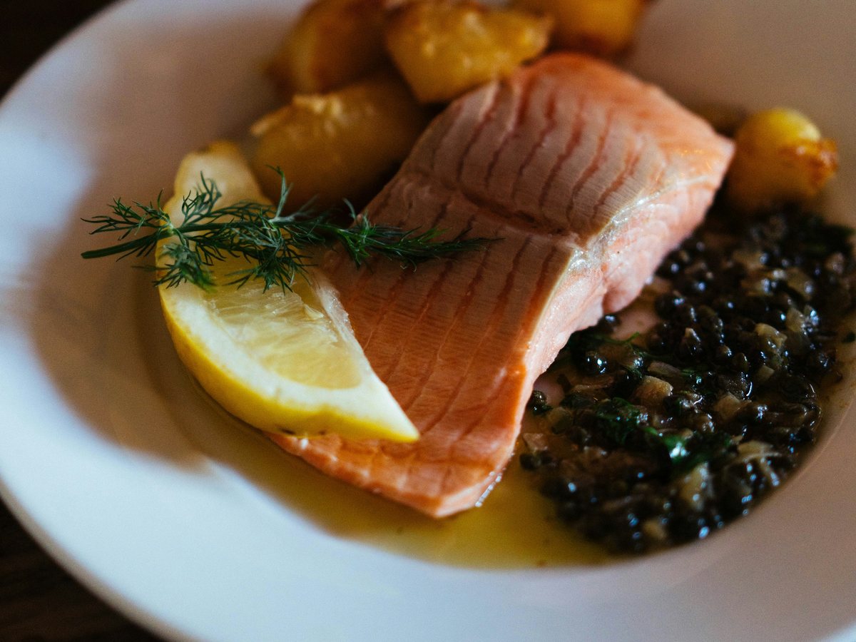 A salmon fillet in a pool of lemon butter with dill, a wedge of lemon and roast potatoes
