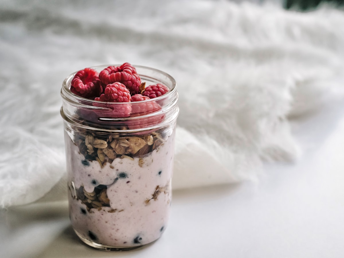 A glass jar of overnight oats layered with yogurt and granola, topped with fresh raspberries