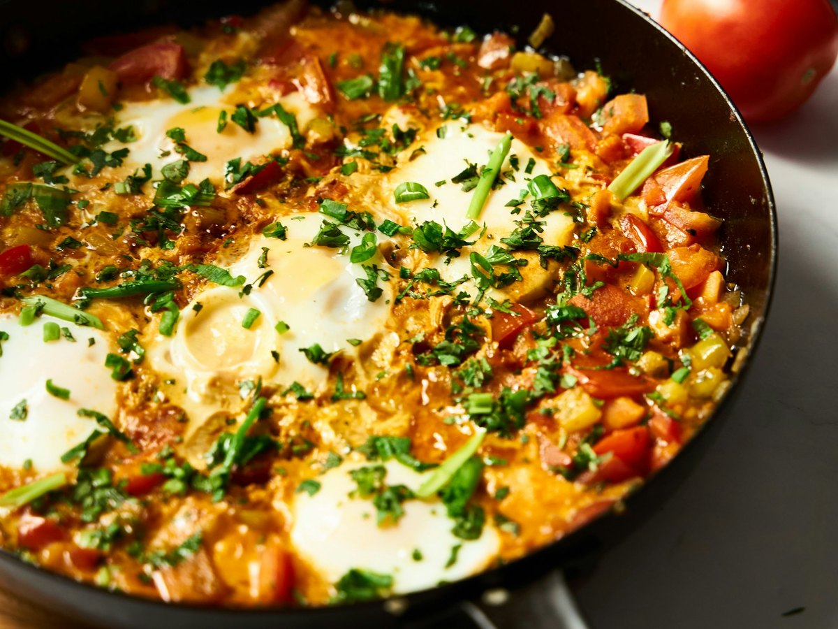 A wide black pan of shakshuka: eggs poached in a thick tomato and pepper sauce, scattered with chopped coriander