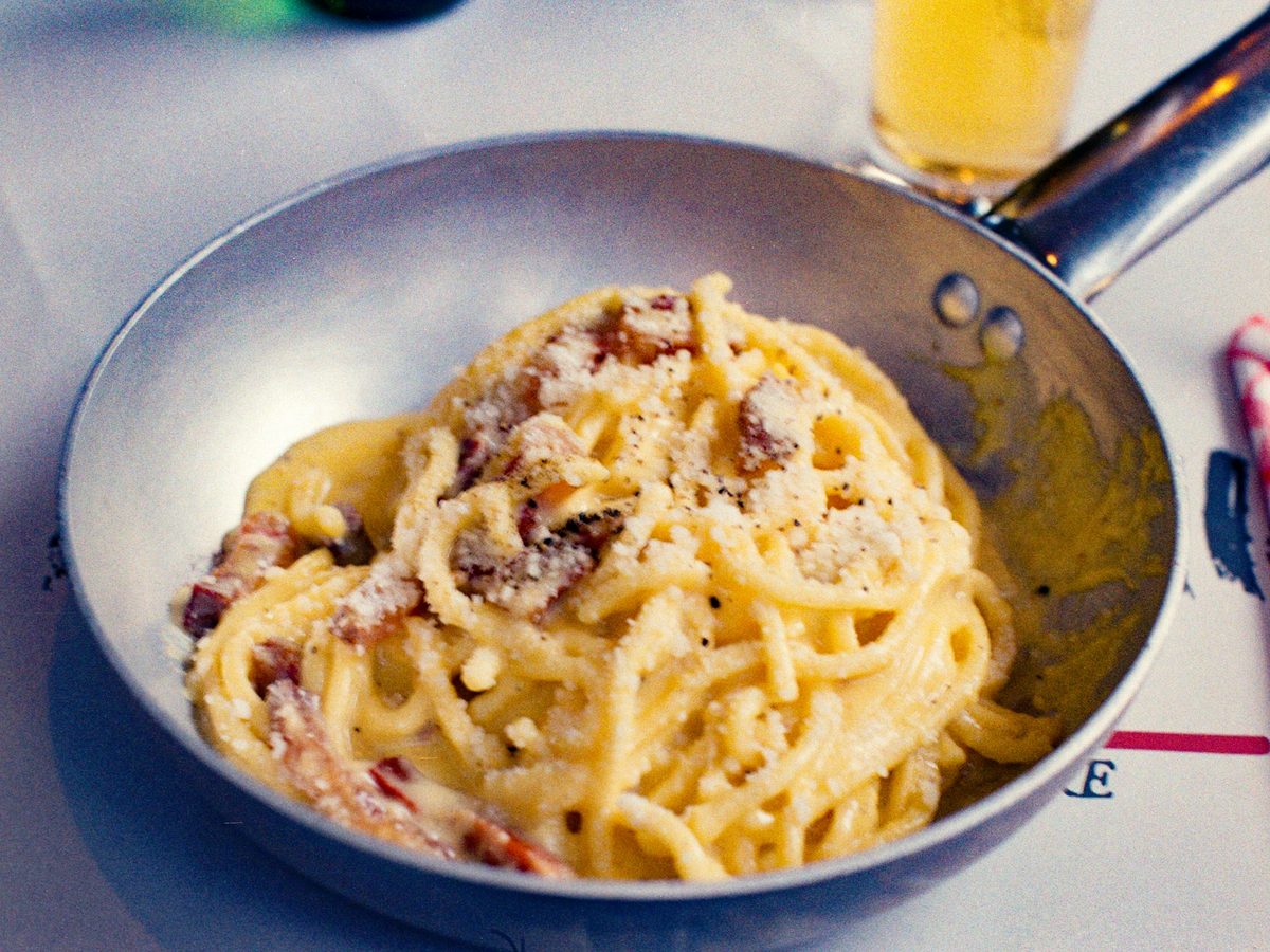 A tangle of spaghetti in a small metal pan, coated in a glossy pale yellow sauce with pieces of crisp guanciale, a heavy scatter of grated pecorino and coarse black pepper